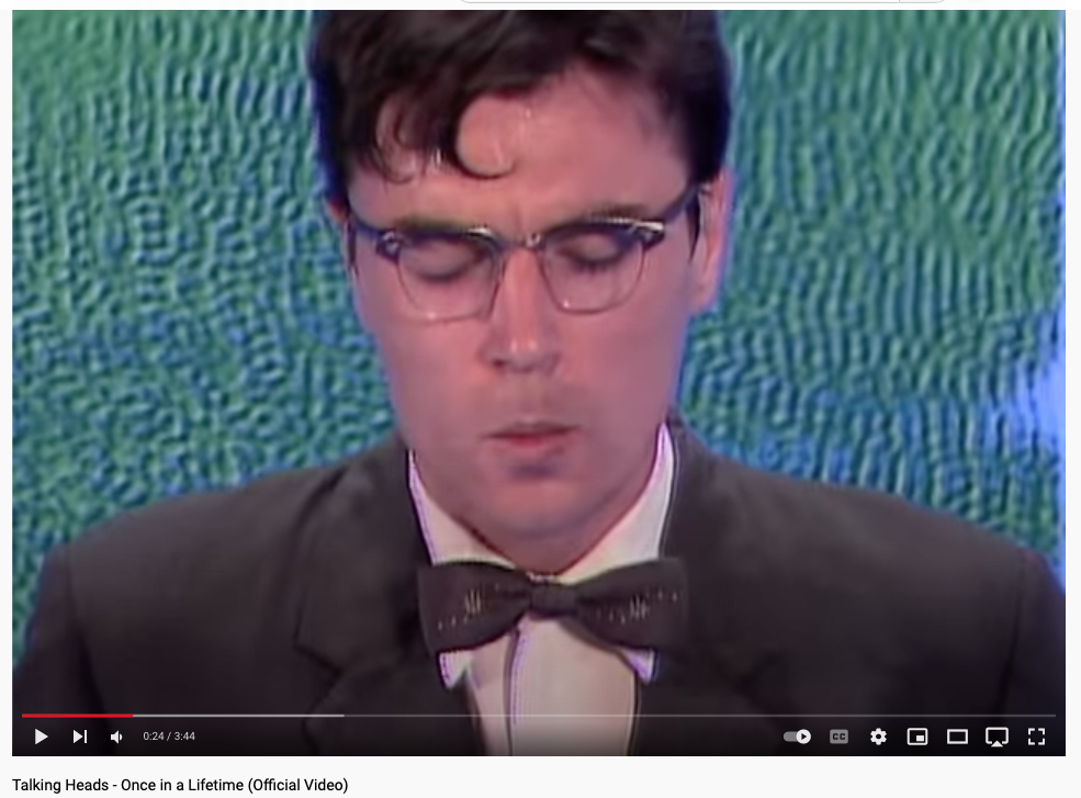 still from Talking Heads "Once In A Lifetime" showing blue-green "water" texture behind David Byrne in suit, glasses, bow tie