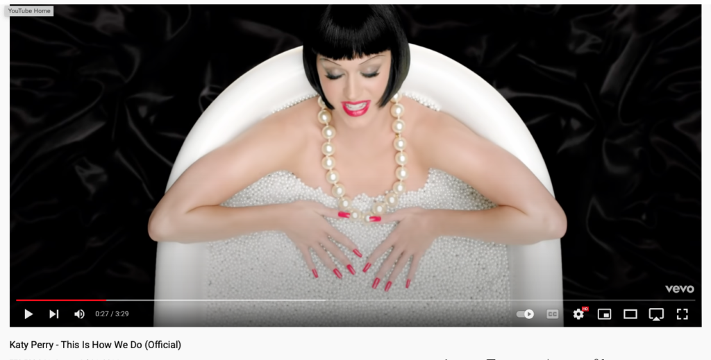 still from Katy Perry "This is how we do" video showing Katy in 1920s bob haircut with giant pearls around her neck, in a bathtub full of pearls, dark rose colored lips and nails